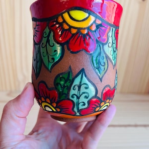 May include: A ceramic cup with a matte brown base featuring a hand-painted floral pattern in vibrant red, yellow, and green. The design includes bold black outlines around the petals and leaves, with a glossy red rim at the top of the vessel.