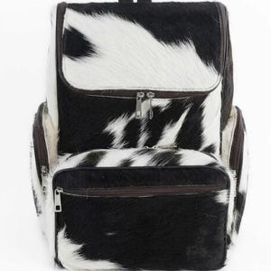 May include: A cowhide backpack with a black and white spotted pattern. The backpack has a top handle, two zippered compartments, and two side pockets. The zippers are silver-toned, and the trim is brown.
