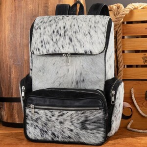 May include: A backpack featuring a white and black speckled animal hide pattern on the main body and front pocket. The bag includes black leather trim, black zippers with silver-toned pulls, and black shoulder straps. The front pocket has a horizontal zipper.