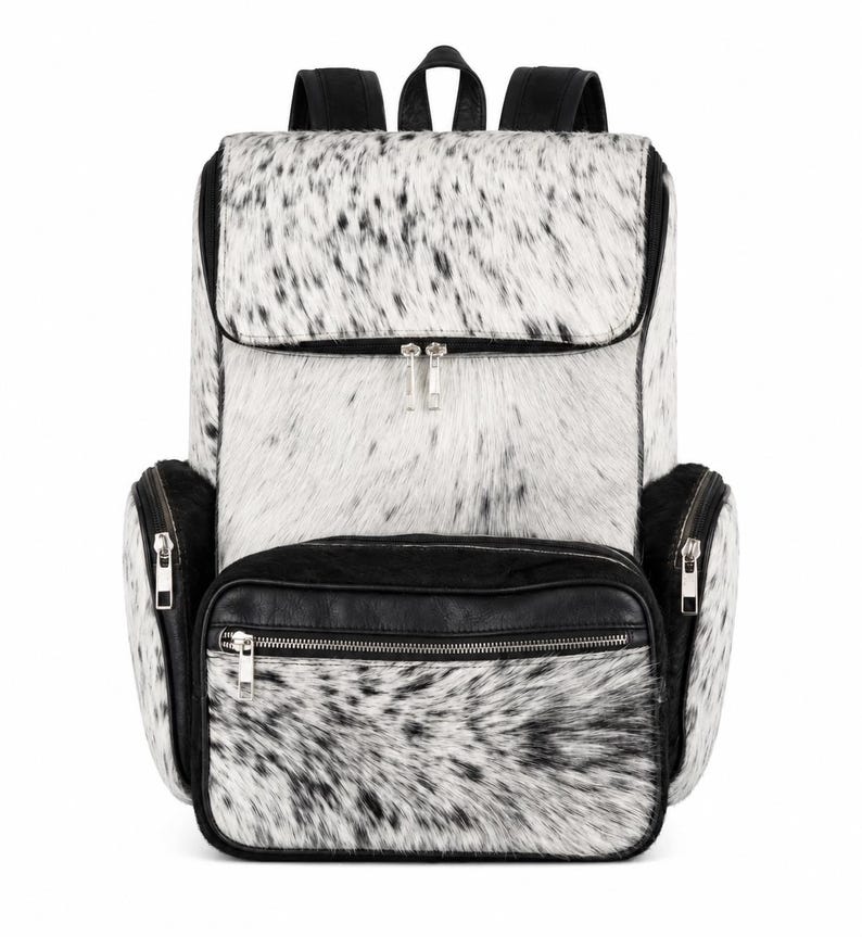 May include: A backpack featuring a white and black speckled animal hair texture on the main body and front pocket. The bag includes black leather trim, silver-tone metal zippers on the front and side compartments, and black shoulder straps.