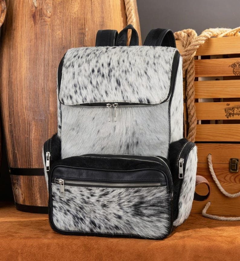 May include: A backpack featuring a white and black speckled animal hide pattern on the main body and front pocket. The bag includes black leather trim, black zippers with silver-toned pulls, and black shoulder straps. The front pocket has a horizontal zipper.