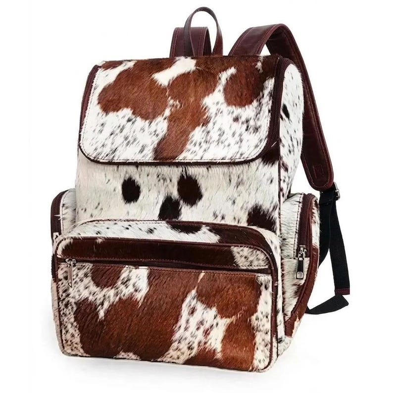 Peut inclure: Un sac &agrave; dos avec un motif de peau de vache marron et blanc et une finition en fourrure textur&eacute;e. Le sac comprend un rabat sup&eacute;rieur, une poche frontale zipp&eacute;e et des bordures en cuir marron fonc&eacute; sur les bords, les bretelles et les fermetures.