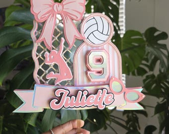 Puede incluir: Adorno para tarta rosa y blanco con temática de voleibol, con el nombre "Juliette" y el número "9". El adorno presenta un balón de voleibol, un lazo y una silueta de jugador de voleibol. El diseño incluye una pancarta y gafas de sol.