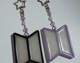 May include: Two matching keyrings. One keyring has a black border, the other has a purple border. Both have a rectangular shape with a silver interior. Each keyring has a star-shaped charm and a string of beads in shades of purple and white.