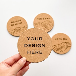 May include: Four round cork coasters with engraved designs and text. One coaster reads "YOUR DESIGN HERE", another "BRECON BEACONS", a third "PEN Y FAN", and the last "CORN DU". The coasters are light brown.