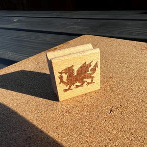 May include: A wooden block featuring a laser-etched dragon design, possibly the Welsh dragon, in brown. The block is made of light-coloured wood and rests on a cork surface. The dragon faces left.