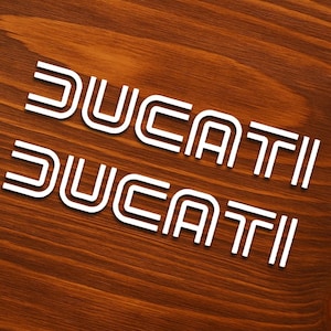 May include: Two white Ducati logos are displayed on a wooden surface. The logos are in a bold, sans-serif font with a double-lined design. The wood has a warm, brown tone with visible grain.