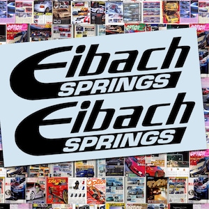 May include: Two black and white Eibach Springs decals on a light blue background, set against a backdrop of car magazine covers. The decals feature the brand name in a stylized font.