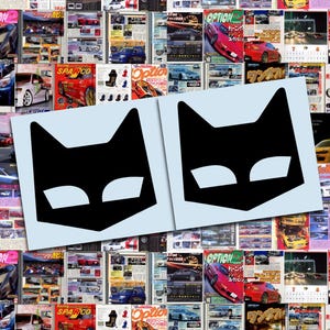 May include: Two black cat-shaped decals with white eye shapes are displayed on a light blue background. The background is a collage of colorful car magazine covers. The decals are a simple, graphic design.