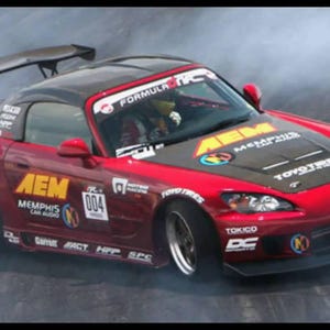 May include: A red Honda sports car with a black hood and spoiler, drifting on a track. The car has sponsor logos including "AEM" and "TOYO TIRES". The number "004" is visible on the side.