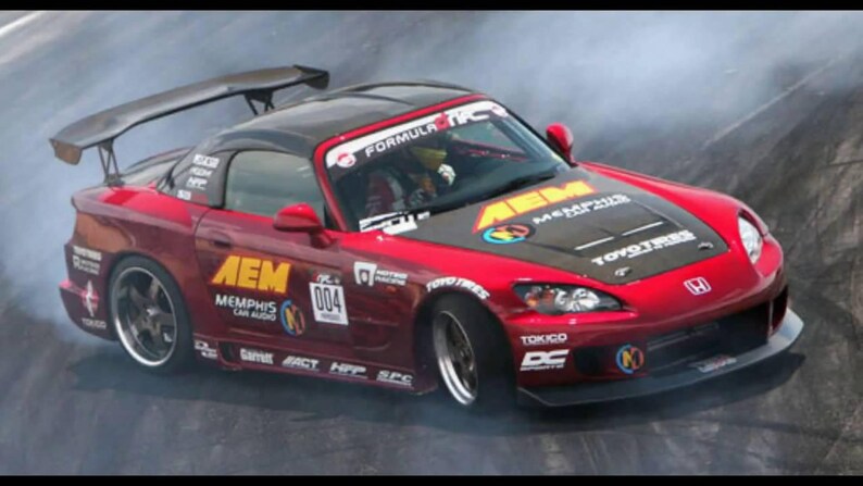 May include: A red Honda sports car with a black hood and spoiler, drifting on a track. The car has sponsor logos including "AEM" and "TOYO TIRES". The number "004" is visible on the side.
