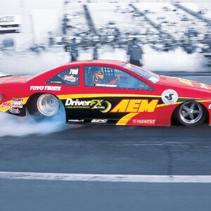 May include: A red race car with black and yellow accents speeds down a track, emitting a cloud of smoke. The car features the logos "DriverFX," "AEM," and "TOYO TIRES." The number 700 is visible on the car's side.