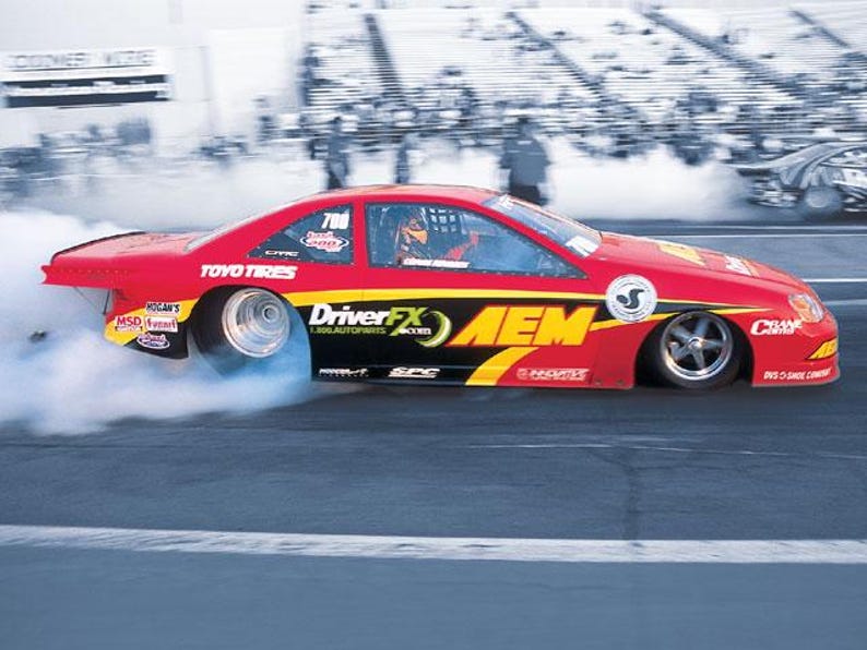 May include: A red race car with black and yellow accents speeds down a track, emitting a cloud of smoke. The car features the logos "DriverFX," "AEM," and "TOYO TIRES." The number 700 is visible on the car's side.