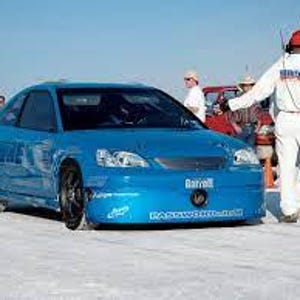 May include: A bright blue sports car with black wheels and a low profile. The car has a streamlined design and is parked on a flat, light-colored surface. Several people in white uniforms and hats are standing nearby.