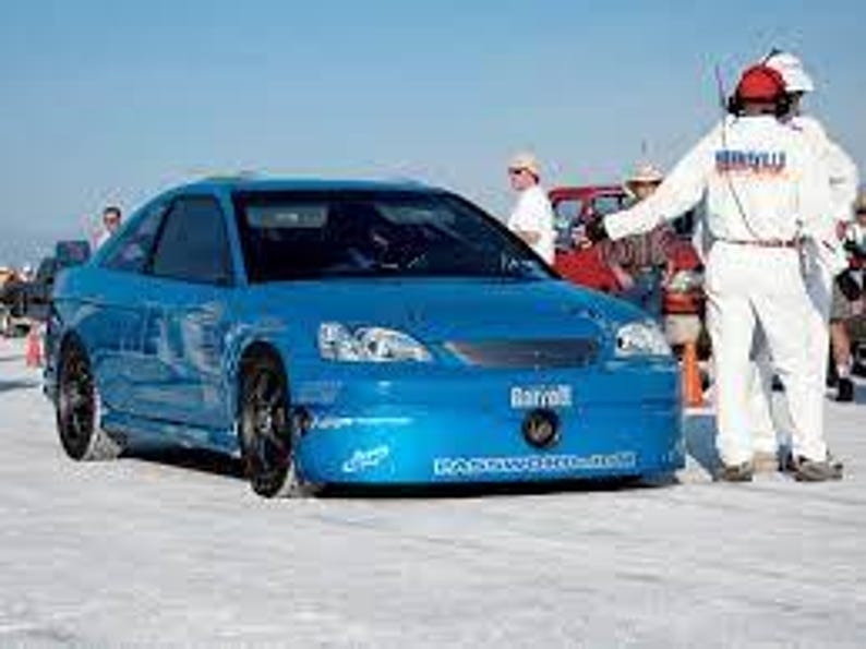 May include: A bright blue sports car with black wheels and a low profile. The car has a streamlined design and is parked on a flat, light-colored surface. Several people in white uniforms and hats are standing nearby.
