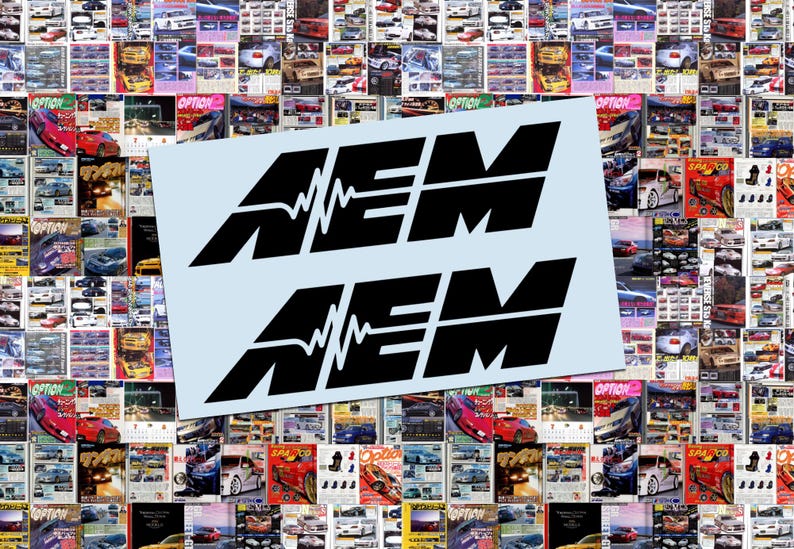 May include: Two black AEM logo stickers on a light blue background, set against a backdrop of car magazine covers. The AEM logo features stylized text with a waveform graphic.