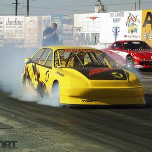 May include: A yellow race car with black accents and sponsor logos is shown on a racetrack, emitting smoke. A red race car is in the background. A person in a black and yellow uniform is standing on the left.