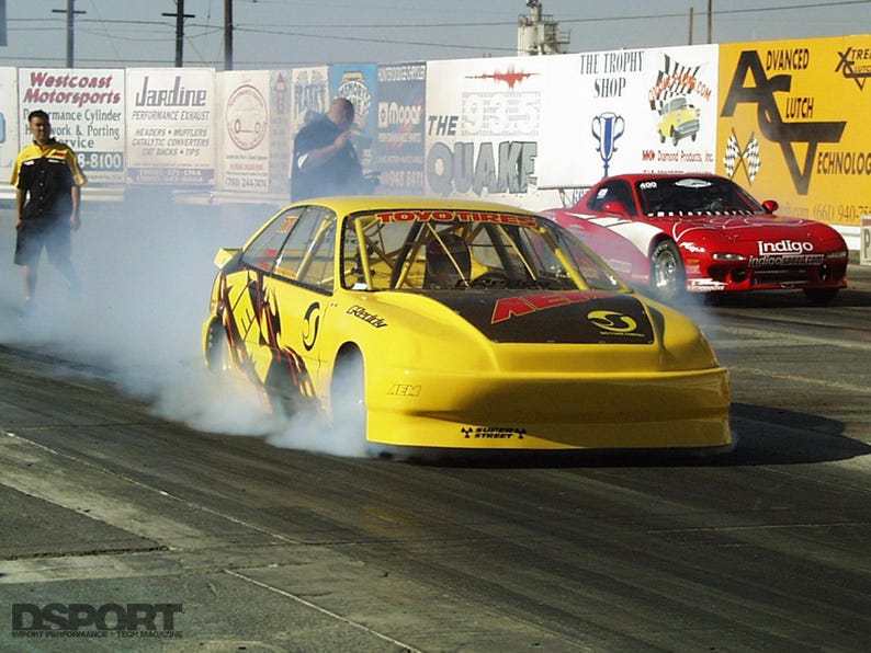 May include: A yellow race car with black accents and sponsor logos is shown on a racetrack, emitting smoke. A red race car is in the background. A person in a black and yellow uniform is standing on the left.