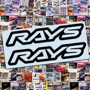 May include: Two black and white "RAYS" logo stickers on a light blue background, set against a collage of car magazine covers. The stickers feature a bold, outlined font, suitable for automotive enthusiasts.