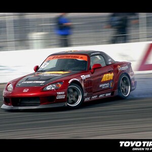 May include: A red Honda sports car drifts on a racetrack, with smoke billowing from the tires. The car has black accents, sponsor decals, and a black roof. The Toyo Tires logo is visible in the bottom right corner.