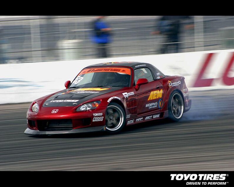 May include: A red Honda sports car drifts on a racetrack, with smoke billowing from the tires. The car has black accents, sponsor decals, and a black roof. The Toyo Tires logo is visible in the bottom right corner.