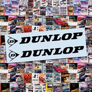 May include: Two black Dunlop logo decals on a light blue background, set against a backdrop of car magazine covers. The Dunlop logo features a stylized 'D' within a triangle. The magazines showcase various car models and automotive content.