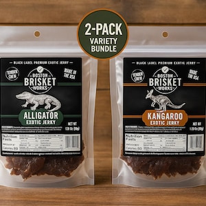Op de afbeelding: Een 2-pack verscheidenheid aan exotische jerky in een doorzichtige plastic verpakking. Eén verpakking bevat alligator jerky, de andere kangoeroe jerky. Elke verpakking heeft een zwart label met de merknaam "Boston Brisket Works" en de tekst "Made in the USA".