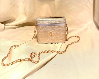 Puede incluir: Un neceser rectangular con acabado texturizado en color blanco roto y el logotipo YSL Beaute en tono dorado en la parte frontal. El bolso cuenta con cierre de cremallera superior, herrajes dorados y una correa de cadena fina y larga.