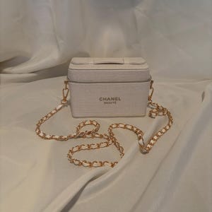 May include: A rectangular off-white vanity case with a textured fabric finish. The front features the text CHANEL BEAUTÉ in dark lettering. It includes a gold-tone metal chain shoulder strap interwoven with off-white material and gold hardware.