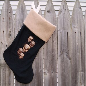 May include: A black Christmas stocking with a tan cuff features an embroidered design of multiple brown skulls.