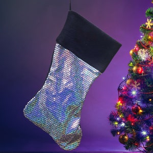 May include: A Christmas stocking with a black cuff and a body covered in iridescent sequins. The stocking hangs against a purple gradient background, with a decorated Christmas tree on the right.