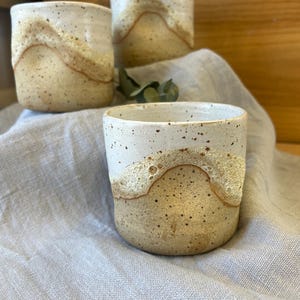 May include: Three handmade ceramic cups. The cups have a speckled, tan base and a creamy white top, separated by a band of textured, light brown glaze. They are arranged on a light grey linen cloth.