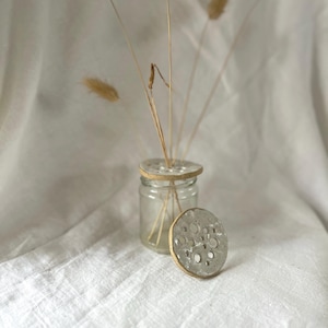 May include: A clear glass jar with a circular, textured, light-coloured ceramic lid featuring multiple holes for flower stems. A second, matching ceramic lid with a similar cratered surface and light-coloured rim rests upright in front of the glass jar.