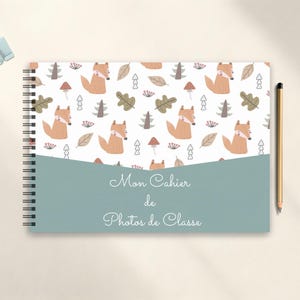 May include: Spiral-bound notebook with a whimsical fox pattern on the cover. The design includes orange foxes, trees, leaves, and mushrooms on a white background. The notebook has a teal-colored band with the text "Mon Cahier de Photos de Classe". A pencil and a binder clip are also visible.