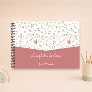 Può includere: Un quaderno a spirale con copertina bianca caratterizzata da un motivo ripetuto di animali del bosco, piante e funghi ad acquerello. La metà inferiore è color rosa antico con la scritta bianca "Les photos de classe d'Anna".