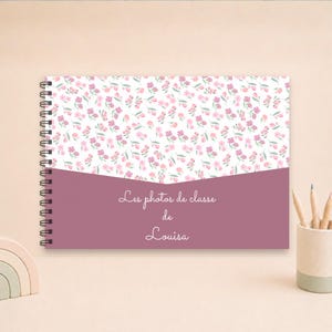 Puede incluir: Un álbum de fotos horizontal con encuadernación en espiral, con una cubierta blanca que presenta un estampado floral rosa y verde en la parte superior y una sección inferior de color malva. El texto "Les photos de classe de Louisa" está en blanco.