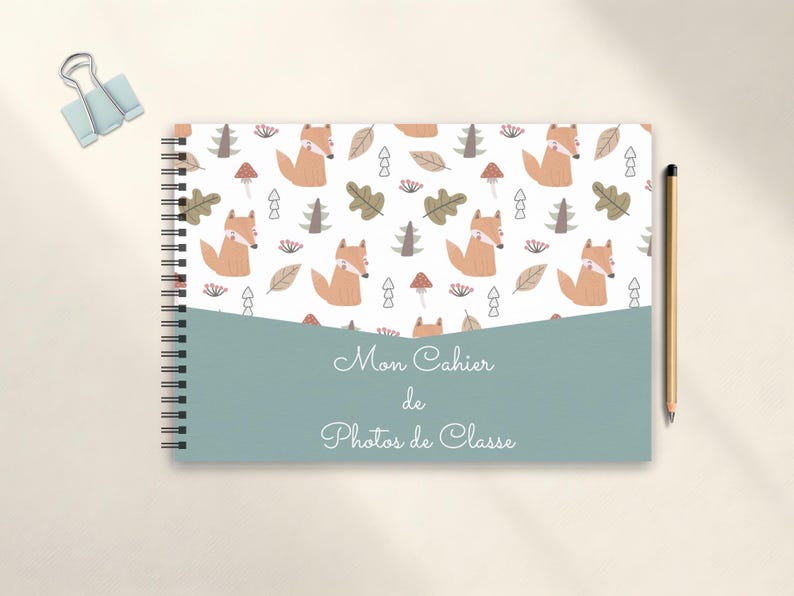 May include: Spiral-bound notebook with a whimsical fox pattern on the cover. The design includes orange foxes, trees, leaves, and mushrooms on a white background. The notebook has a teal-colored band with the text "Mon Cahier de Photos de Classe". A pencil and a binder clip are also visible.