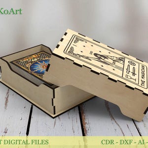 Può includere: Una scatola di legno color chiaro per carte dei tarocchi con un coperchio con un disegno tagliato al laser della carta dei tarocchi "Il Mago". La scatola è aperta, rivelando un mazzo di carte con un disegno blu e oro.