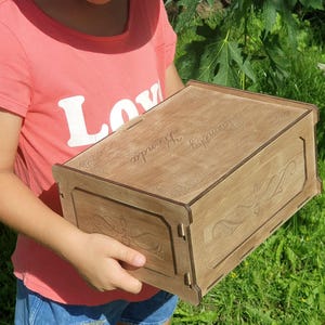 Puede incluir: Una caja de madera marrón claro con tapa con bisagras y tallas florales decorativas. El nombre "Amanda" está grabado en la parte superior. La caja está sostenida por una persona que lleva una camiseta rosa.