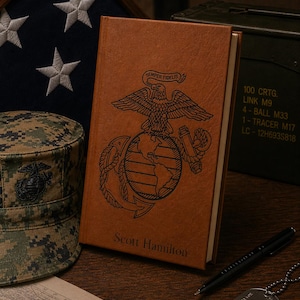 Könnte beinhalten: Ein braunes, ledergebundenes Notizbuch mit dem Emblem des US Marine Corps und dem Text "Semper Fidelis". Der Name "Scott Hamilton" ist unten eingraviert. Ein Stift, eine Erkennungsmarke und eine Militärkappe sind ebenfalls zu sehen.