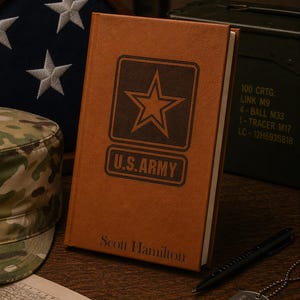 May include: A brown leather-bound journal with the U.S. Army logo and the name "Scott Hamilton" embossed on the cover. A black pen and dog tags are next to the journal. An American flag and a military hat are in the background.
