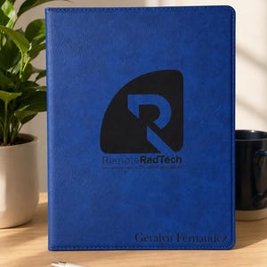 May include: A royal blue leather-textured portfolio featuring a black logo with the text RemoteRadTech and the subtitle Your remote team of CT and MRI technologists. The name Geralyn Fernandez is printed in black at the bottom, with a matching pen.