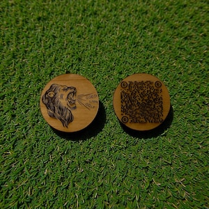 Peut inclure: Deux marqueurs de balle de golf en bois ronds. L'un représente un lion rugissant avec le texte "GOOD LION GOLF". L'autre a un code QR. Ils sont posés sur une surface verte.