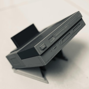 May include: A gray Sony PlayStation 1 console replica, angled on a stand. The console features a disc drive, power button, and various ports. The stand is black and supports the console at an angle, showcasing the side profile.