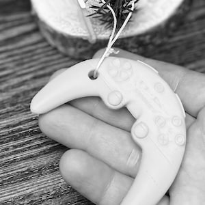 Op de afbeelding: Een crèmekleurig, halvemaanvormig ornament dat lijkt op een videogamecontroller. Het ornament heeft knoppen en richtingspad details. Er is een touwtje bevestigd om op te hangen. Het ornament wordt in een hand gehouden, met een kleine tak groen en een houten schijf op de achtergrond.