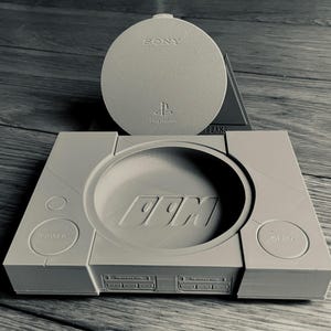 Puede incluir: Réplica gris claro de una consola PlayStation 1 con una cubierta de disco a juego. La consola presenta el texto "99M" y los botones "POWER" y "OPEN". La cubierta del disco está grabada con "SONY" y el logotipo de PlayStation.