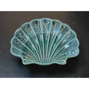May include: A ceramic bowl shaped like a large seashell with a scalloped edge. The bowl features a glossy glaze in shades of teal, blue, and green, with a mottled, crystalline texture concentrated along the upper ridges and a smooth finish.