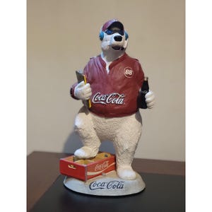 May include: A white polar bear figurine wearing a maroon jacket with the number 88 and a Coca-Cola logo, a cap, and blue headphones. The bear holds a clipboard and a small bottle, standing on a red Coca-Cola crate base with the Coca-Cola logo.
