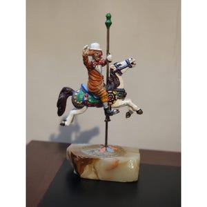 May include: A carousel figurine featuring a clown in a brown suit riding a white horse with a dark mane. The horse has a green saddle with red and blue accents. The figure is mounted on a thin metal pole with a green finial atop a stone base.
