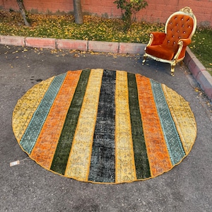 Puede incluir: Una alfombra redonda con rayas verticales en tonos amarillo, naranja, verde, turquesa y negro. La alfombra tiene un aspecto envejecido y vintage. La alfombra está colocada sobre una superficie de asfalto.